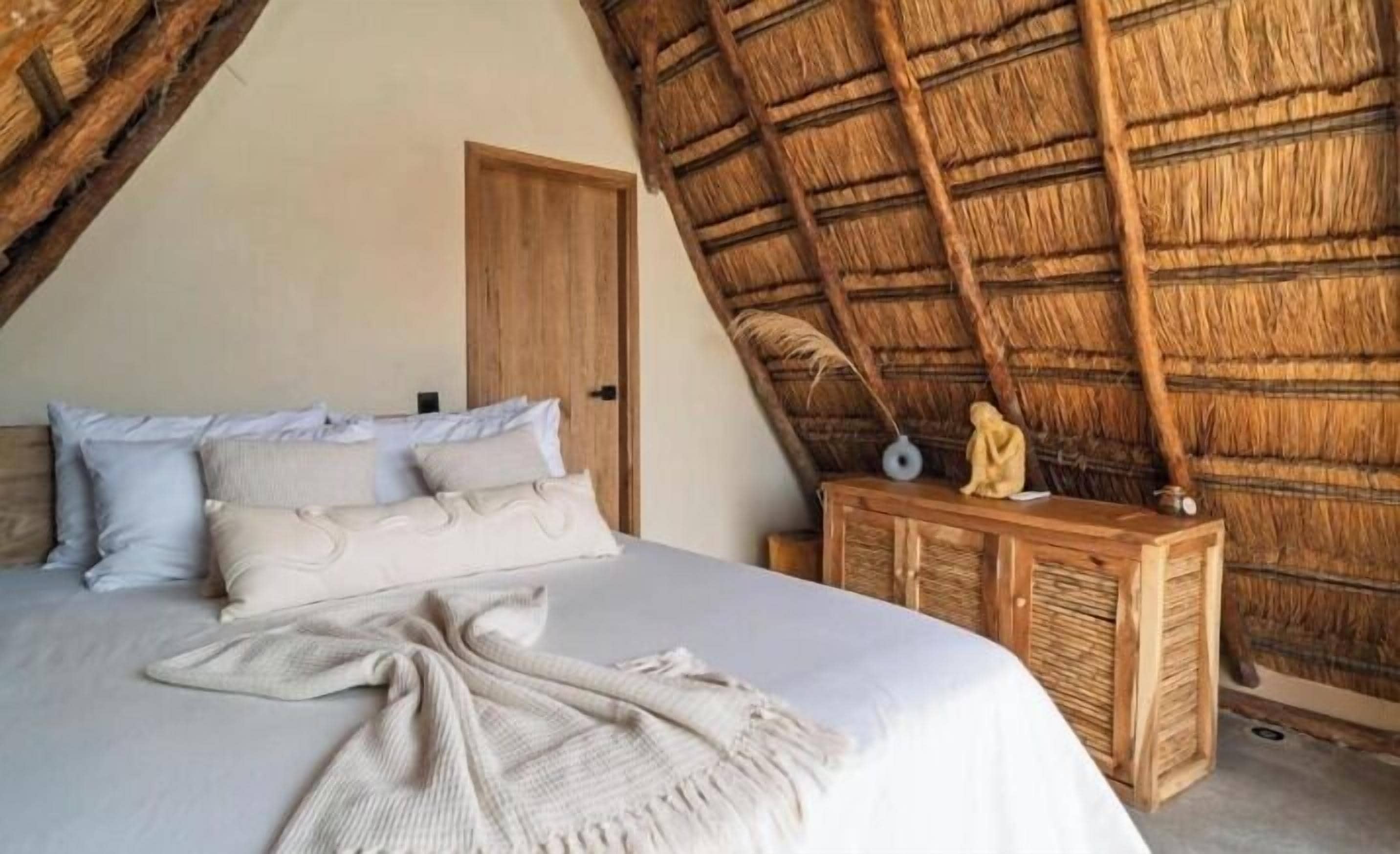 A bedroom with a bed, pillows, a blanket, a wooden nightstand, and a thatched ceiling.