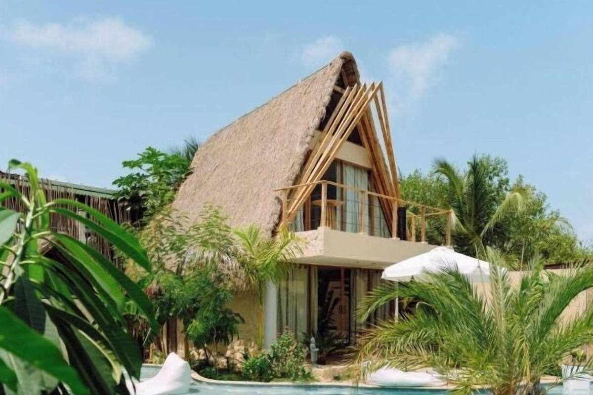 Un resort con un edificio de techo de paja, piscina y vegetación tropical.