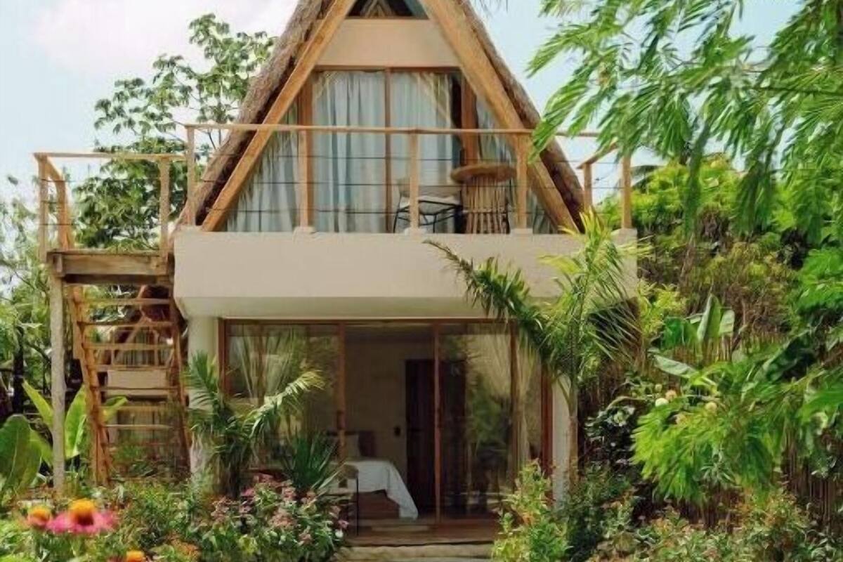 Una casa de dos pisos con techo de paja, rodeada de abundante vegetación y una piscina.