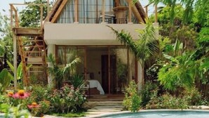 Una casa de dos pisos con techo de paja, rodeada de exuberante vegetación y una piscina.