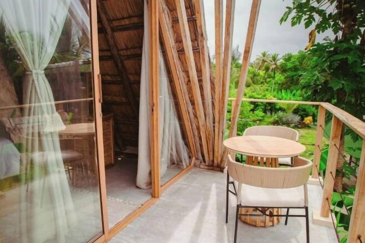 Un balcón con mesa y sillas de madera, una puerta de cristal con cortinas y vistas a zonas verdes.