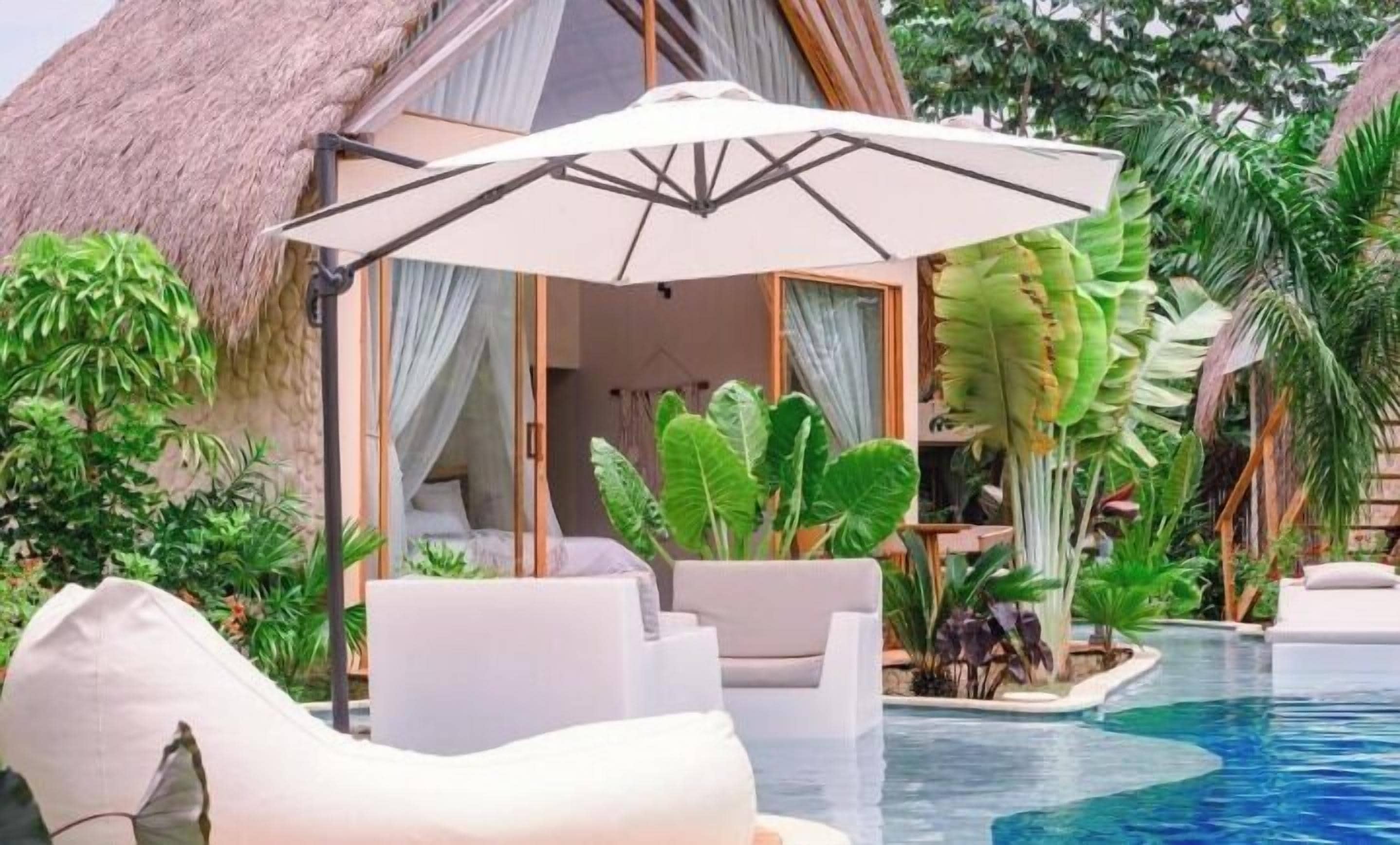 Un área junto a la piscina con una estructura de techo de paja, sillas de descanso y plantas tropicales.