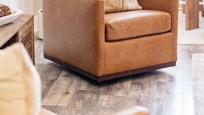 Un sofá de cuero marrón con un cojín, una mesita de madera y una planta en maceta en un salón moderno con suelo de madera y un comedor al fondo.