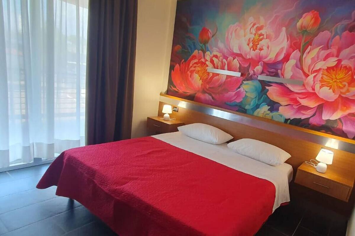 A hotel room with a large bed, white pillows, a red bedspread, and a floral painting on the wall.