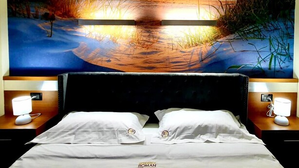 A hotel room with a large wall mural of a sunset over water, a double bed with white linens, and bedside lamps. - Residence Roman (Pomezia)