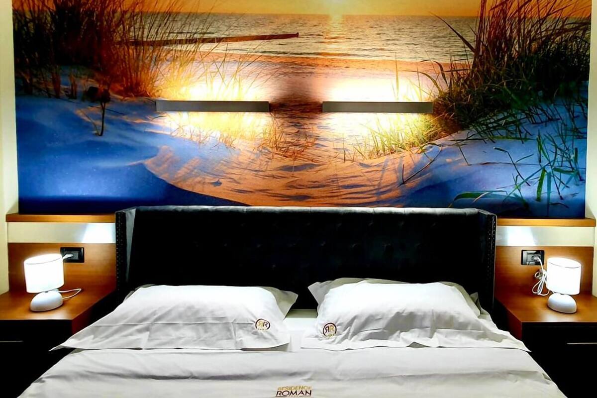 A hotel room with a large wall mural of a sunset over water, a double bed with white linens, and bedside lamps.