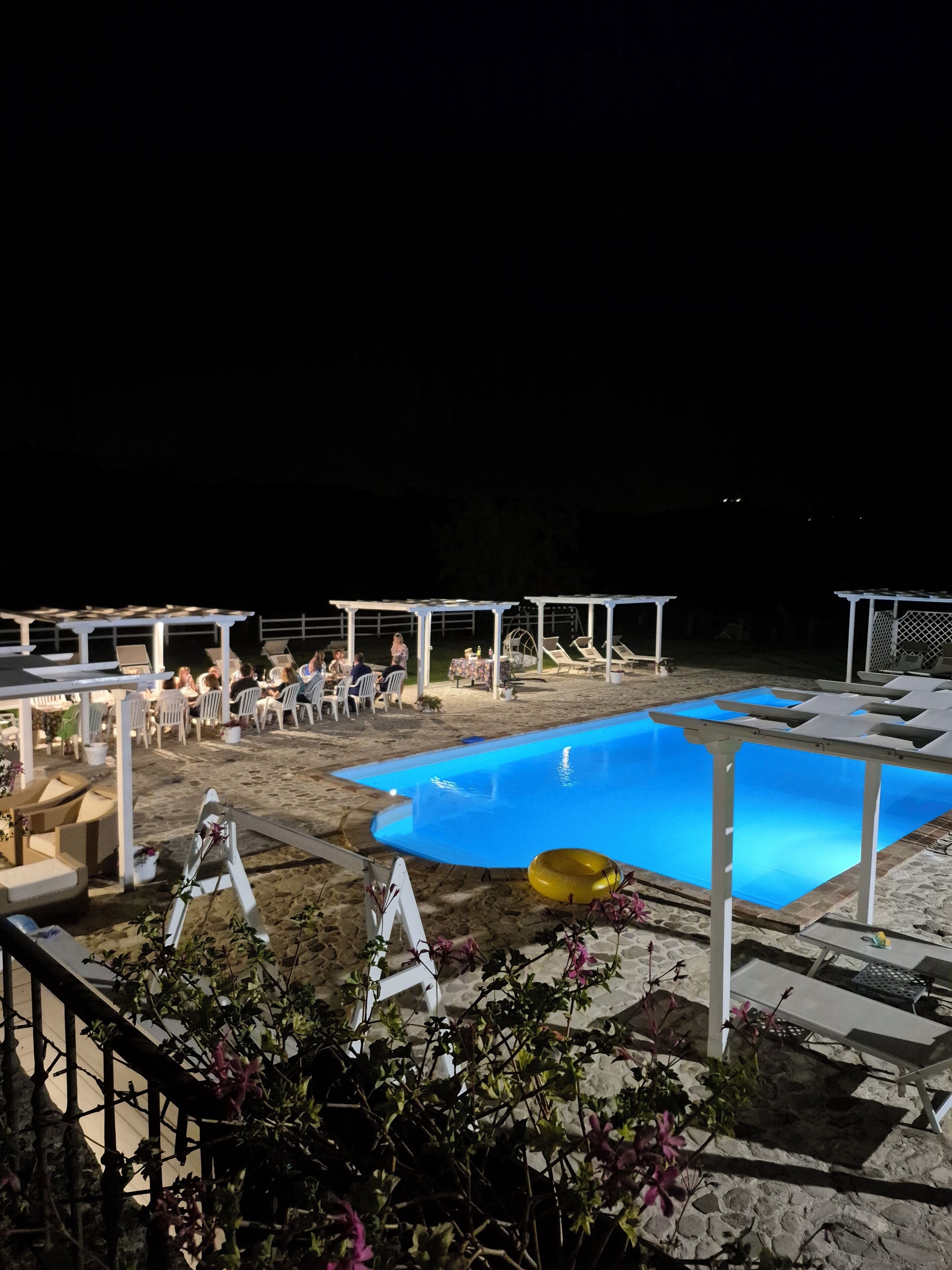 Un'area piscina con una piscina blu, poltroncine bianche e tavoli sotto pergole.