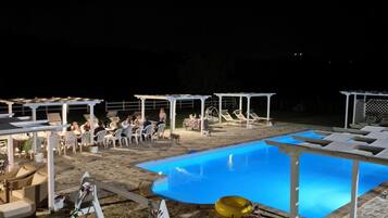 Un'area piscina con una piscina blu, poltroncine bianche e tavoli sotto pergole.
