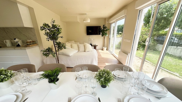 Un espace repas moderne avec une table blanche dressée pour six personnes, entourée de chaises. On aperçoit un lit avec une literie blanche à l’arrière-plan, et un téléviseur est fixé au mur.