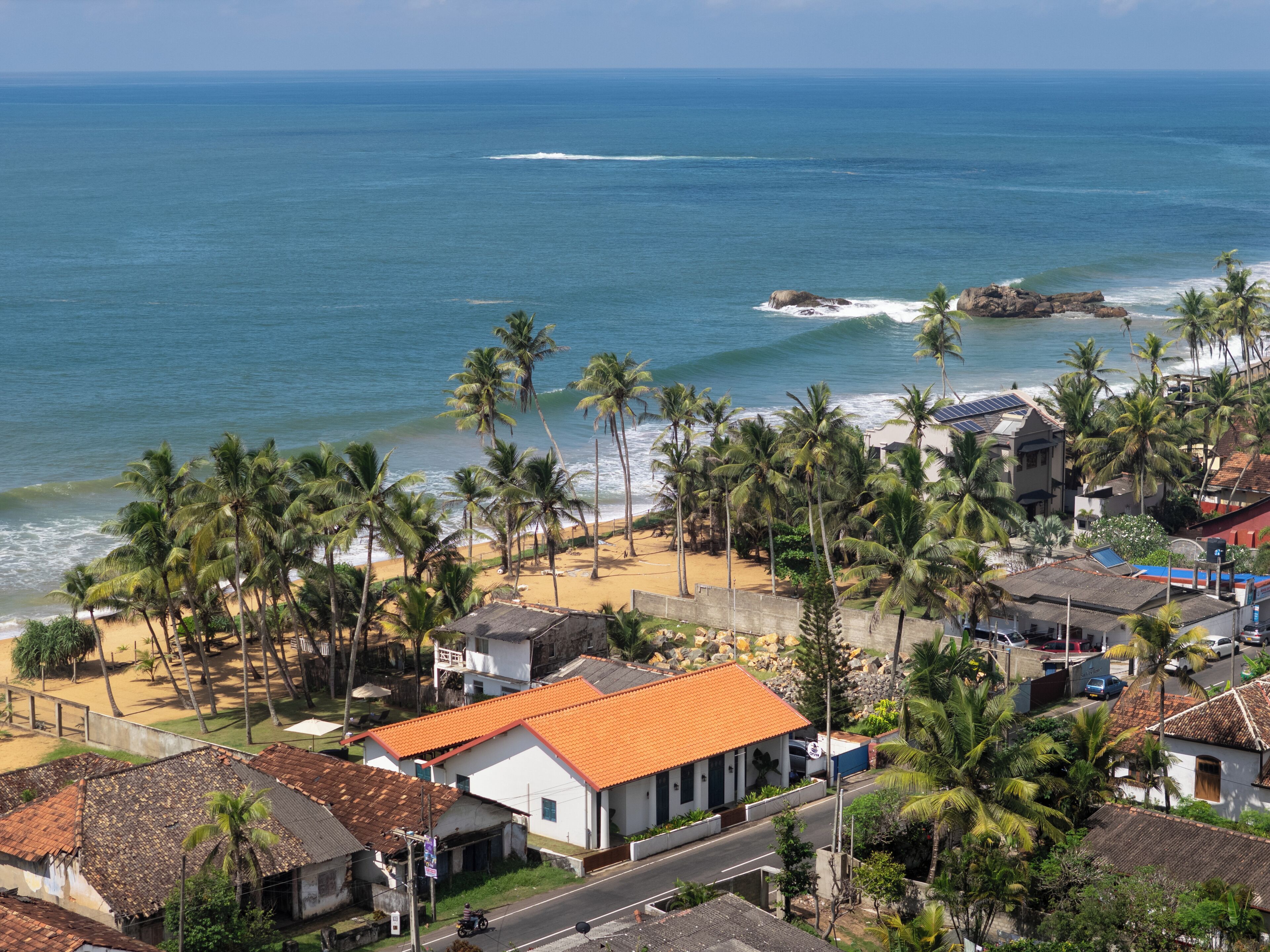 Un pueblo costero con playa, palmeras y casas con techos de tejas rojas.