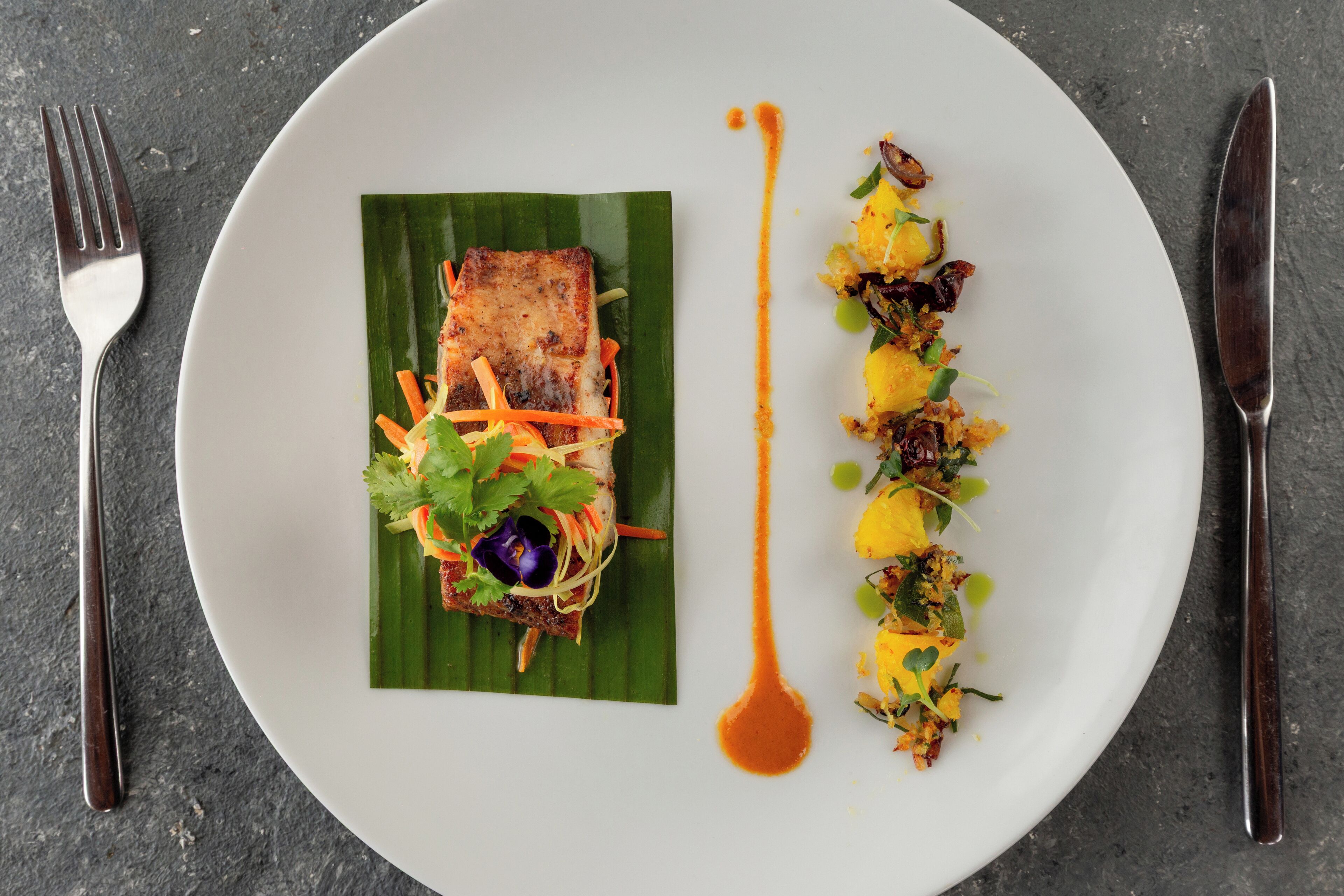A white plate with a piece of grilled fish on a banana leaf, accompanied by a side of vegetables and a drizzle of sauce.