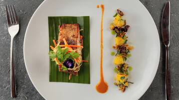 A white plate with a piece of grilled fish on a banana leaf, accompanied by a side of vegetables and a drizzle of sauce.