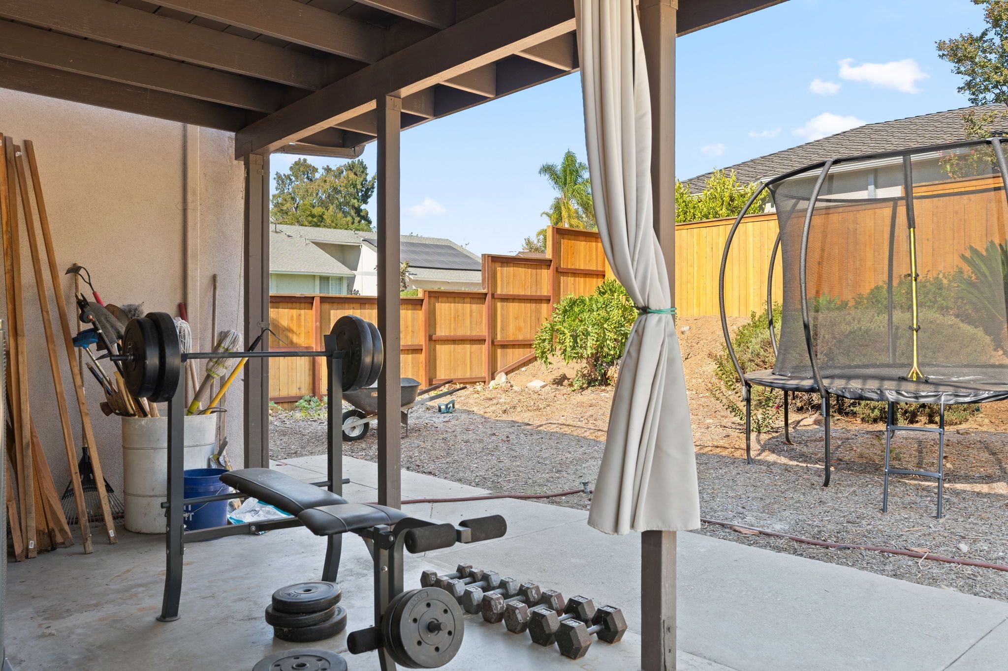 A covered outdoor gym area with exercise equipment, a trampoline, and a wooden fence in the background.