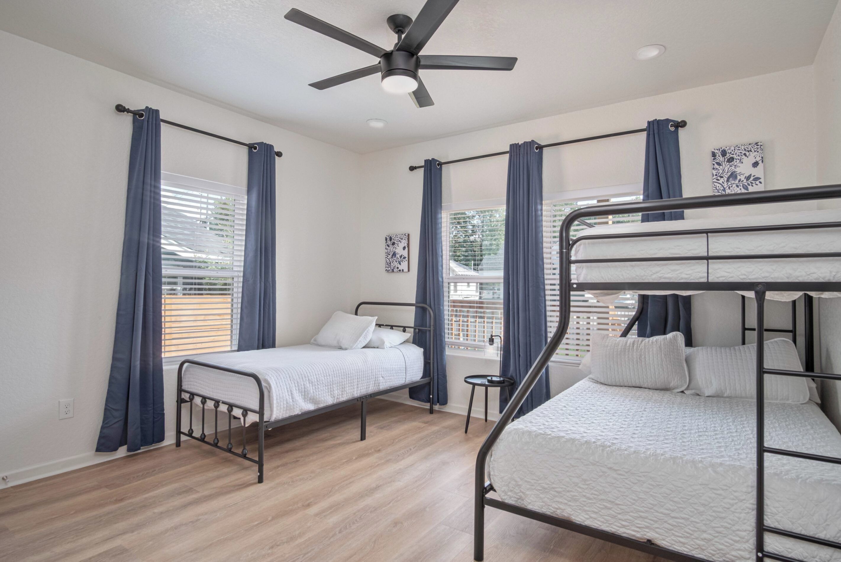 Une chambre avec deux lits simples et un lit superposé, un plancher en bois, un ventilateur de plafond et des fenêtres avec des rideaux.