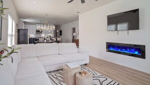 Una sala de estar moderna con un sofá modular, una chimenea, un ventilador de techo y una cocina al fondo.