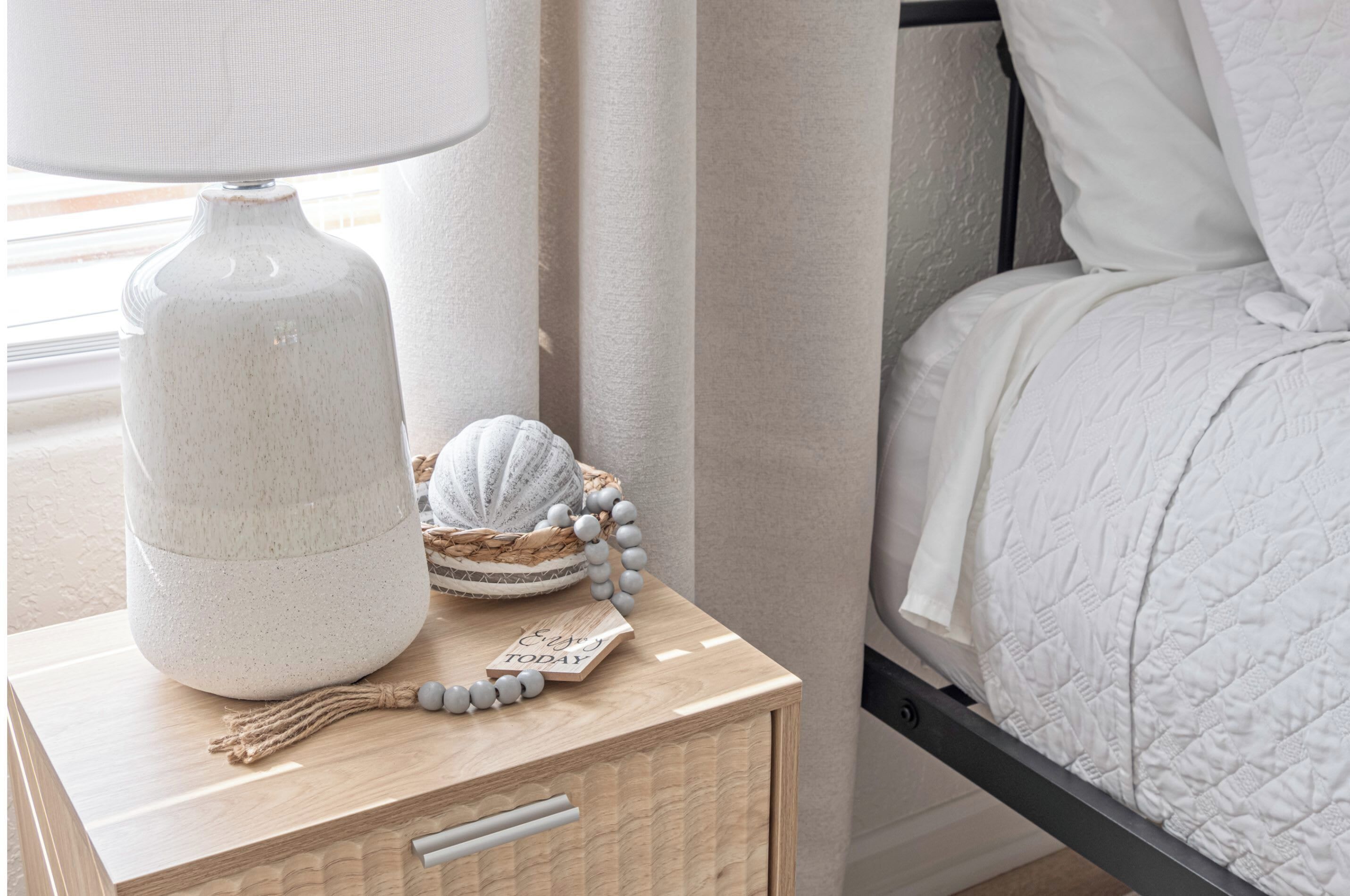 A neatly made bed with a white quilt, a bedside lamp, a small wooden nightstand with a decorative shell and a beaded string, and a framed picture on the wall.