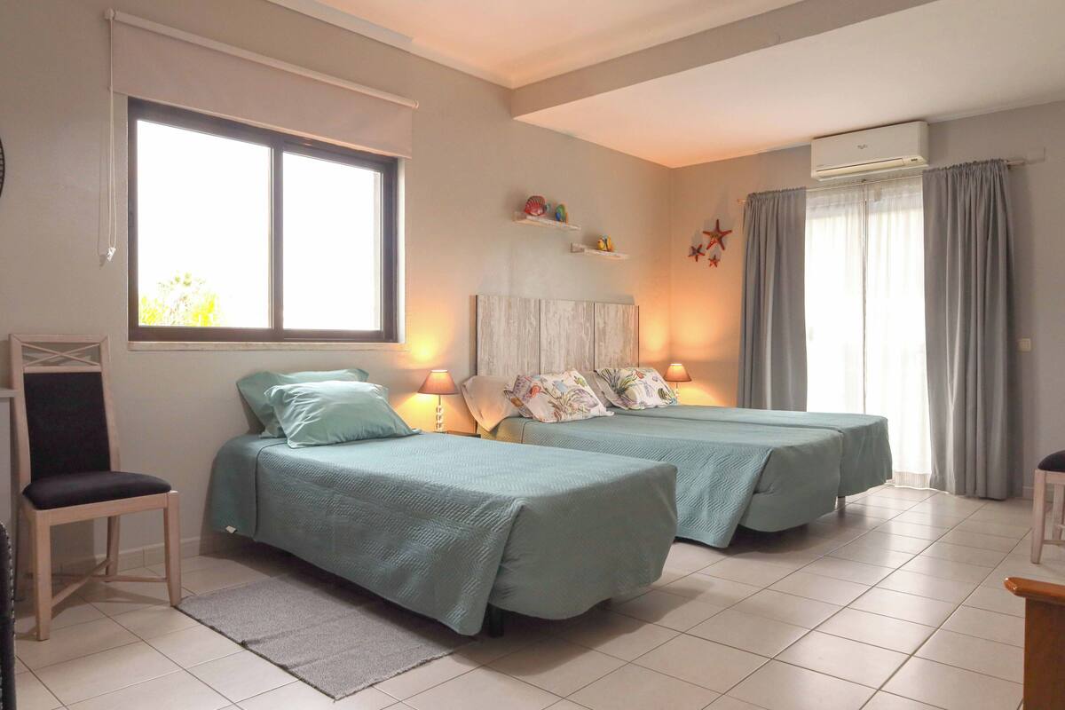 Ein Schlafzimmer mit zwei Betten, einem Sessel, einer Kommode mit Dekoration und einem Fenster mit Vorhängen.