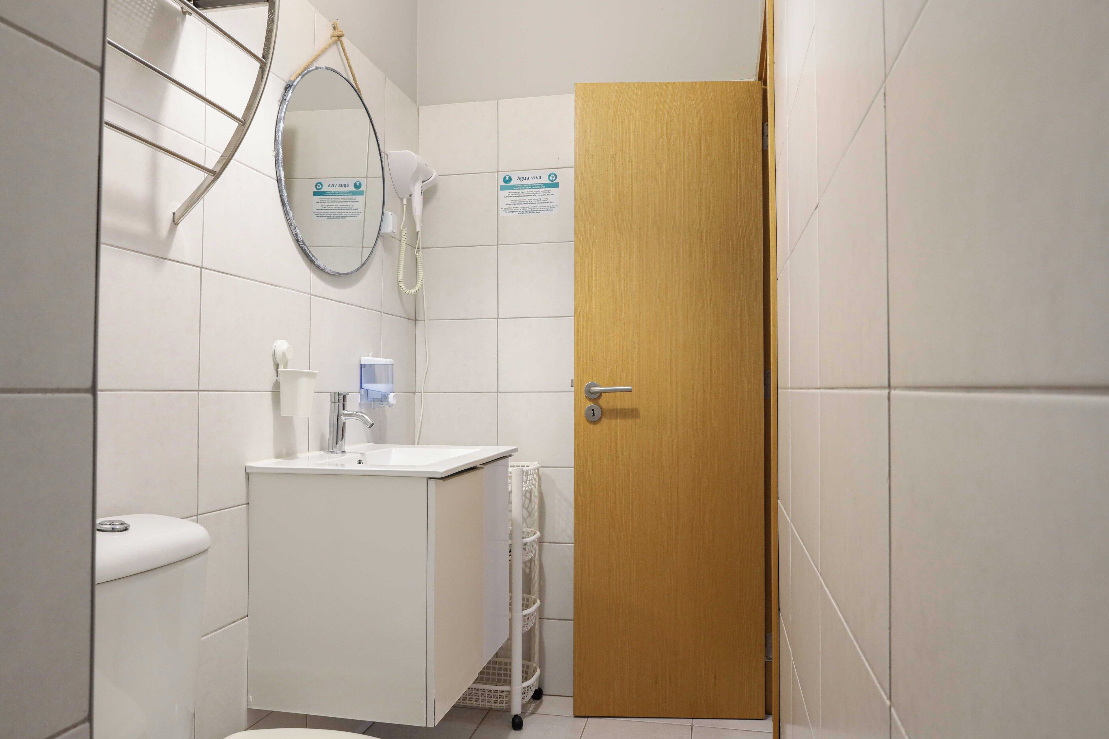 Een badkamer met een houten deur, witte tegels, een wastafel, een spiegel en een handdoekhouder.