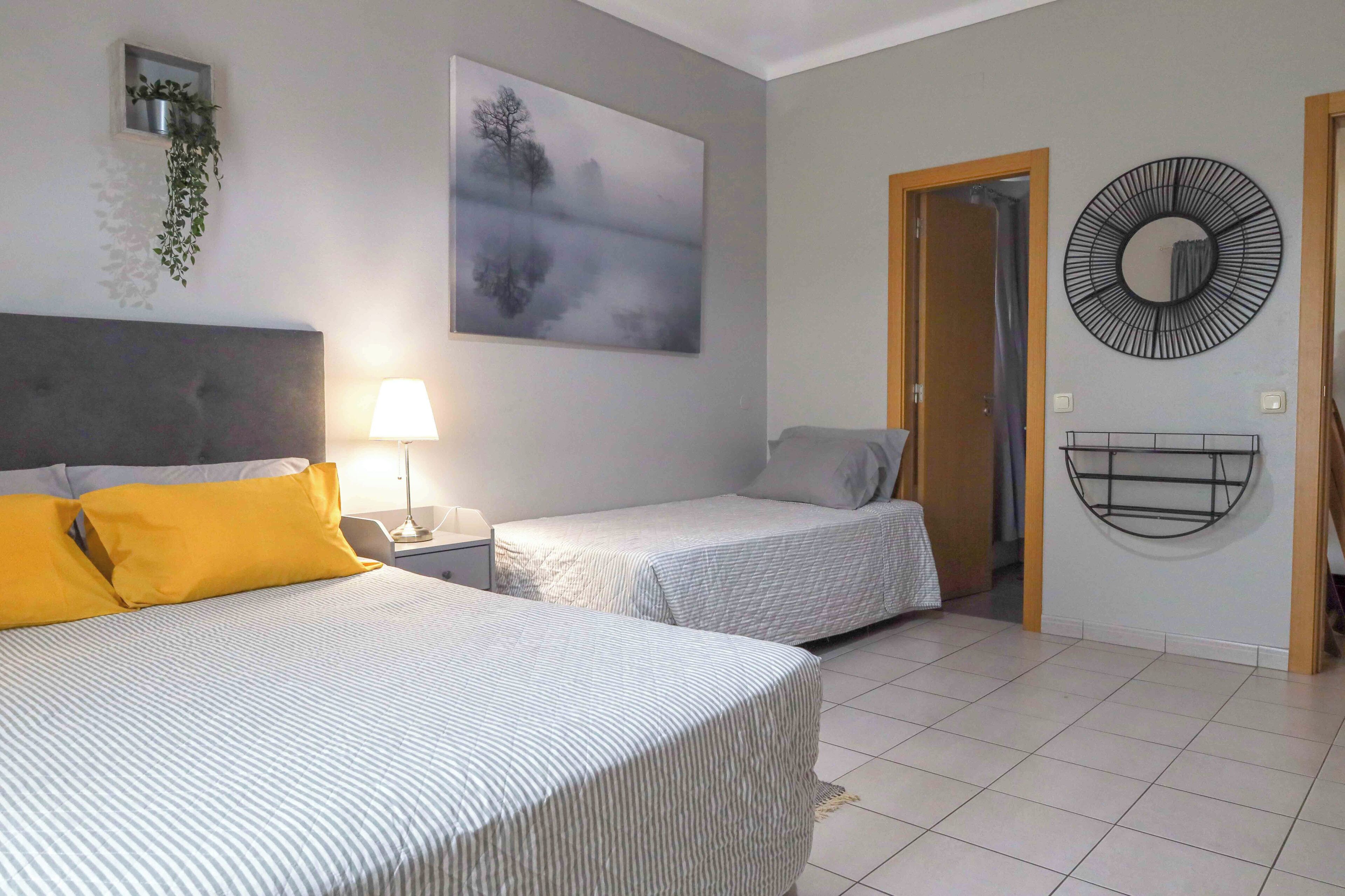 A hotel room with two beds, a nightstand, a wall-mounted plant, a painting, a circular mirror, and a door leading to another room.