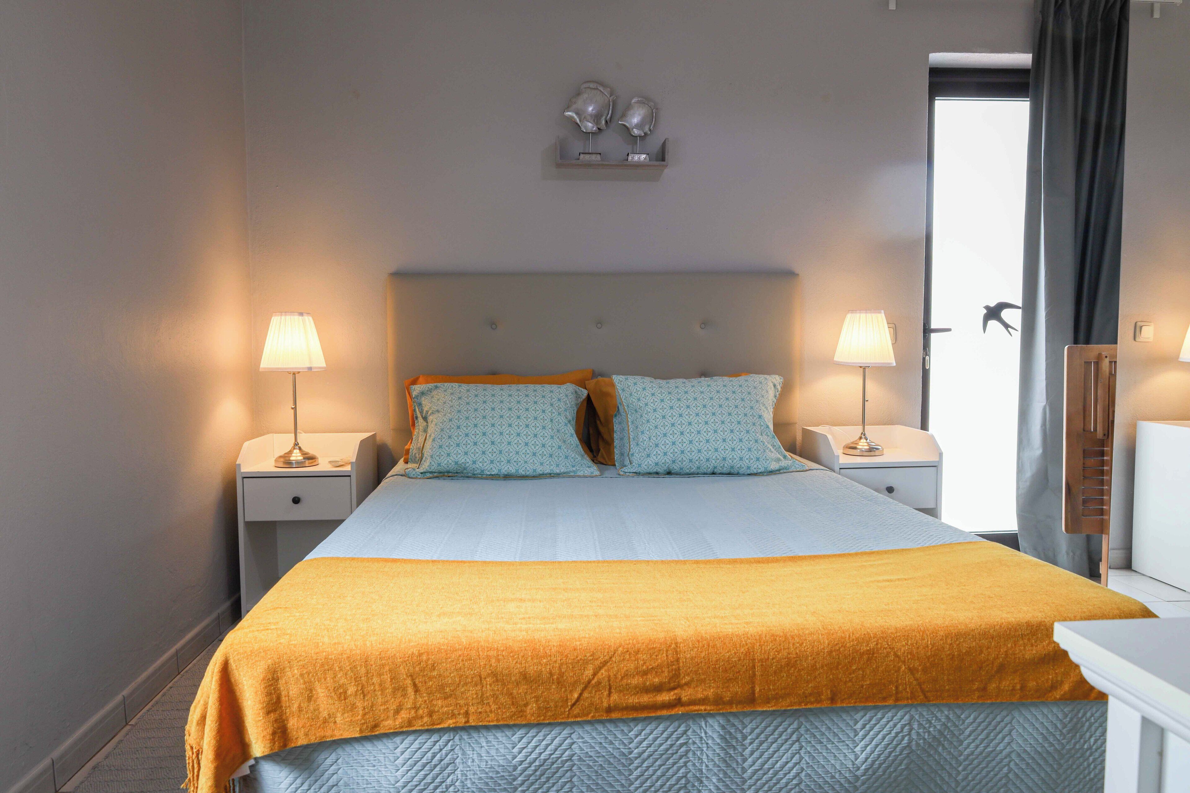 A hotel room with a bed, two bedside lamps, a nightstand, a dresser, and a door with a view.