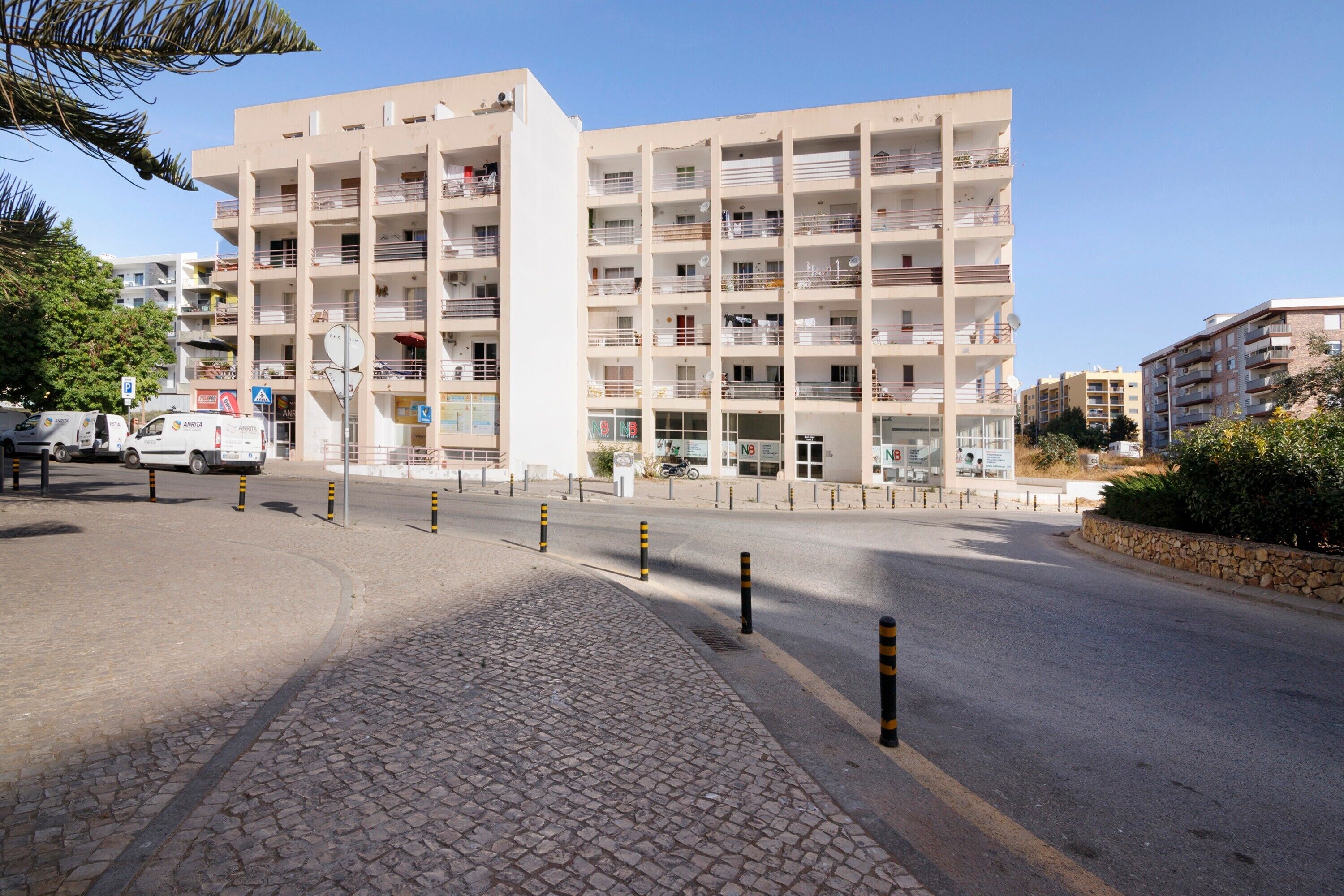 Un edificio residencial de varios pisos con balcones, una calle pavimentada, pilares de seguridad y cielo despejado.