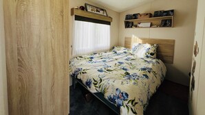 Ein Schlafzimmer mit einem Bett, einem Holz-Kopfteil, einem Regal mit Dekoration und einem Fenster mit leichtem Vorhang.