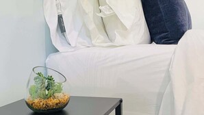 A bedroom with a bed, white bedding, a dark blue pillow, and a small table with a glass bowl containing plants. - Centrally + Conveniently Located (Providence)