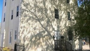A two-story building with a white exterior, a black gate, and a tree casting shadows on the building. - Centrally + Conveniently Located (Providence)