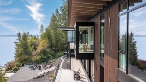 En moderne bygning med glasfacade, udsigt over en sø, udendørs siddeområde og en træterrasse.
