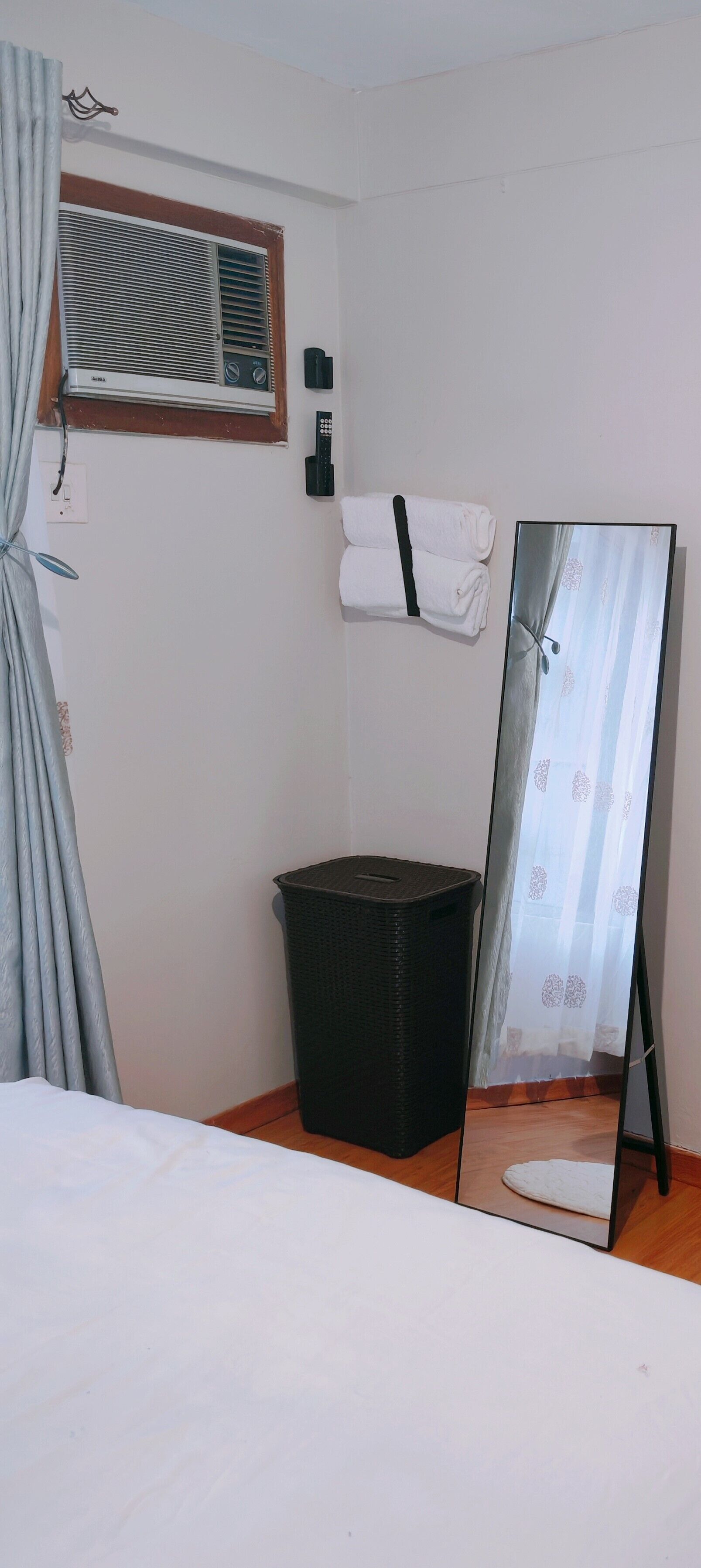 Una habitación con cama, un espejo de pie, un cubo de basura, aire acondicionado instalado en la pared y una ventana con persianas.
