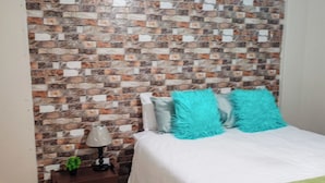 Ein Schlafzimmer mit einem Kopfende aus Ziegelsteinen, einem Bett mit weißer Bettwäsche und türkisfarbenen Kissen, einem kleinen Tisch mit einer Lampe und einer grünen Tagesdecke.