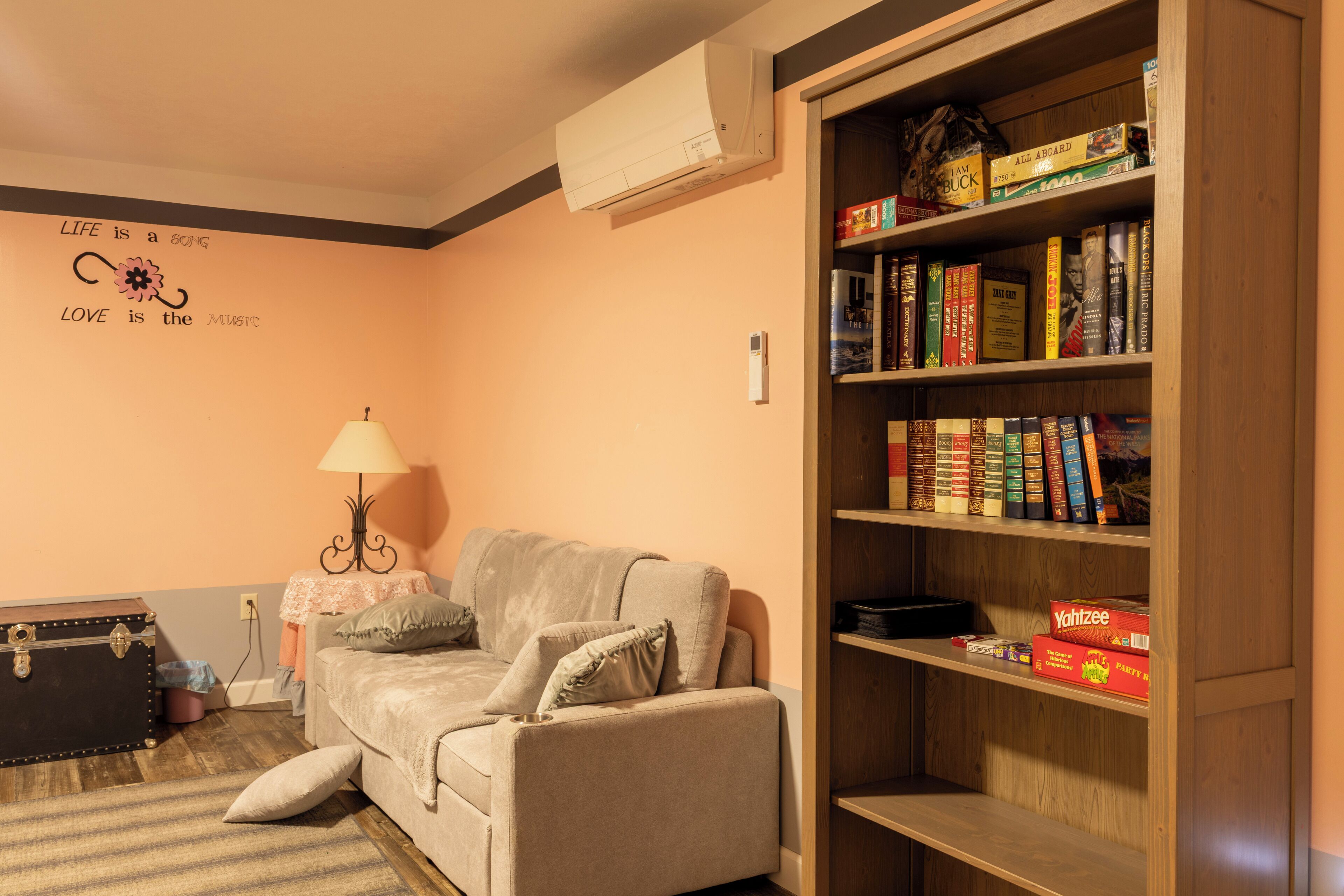 Un salon chaleureux avec un canapé, une lampe, une bibliothèque remplie de livres et un mur orné d’une citation inspirante.