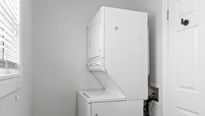 A compact laundry room with a stacked washer and dryer, a window with blinds, and a wooden floor. - Bright 2BR Loft WiFi Parking (St. John's)