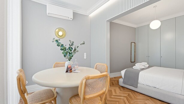 A modern room with a round dining table, four wicker chairs, a bed with a grey duvet, and a wall-mounted air conditioner.
