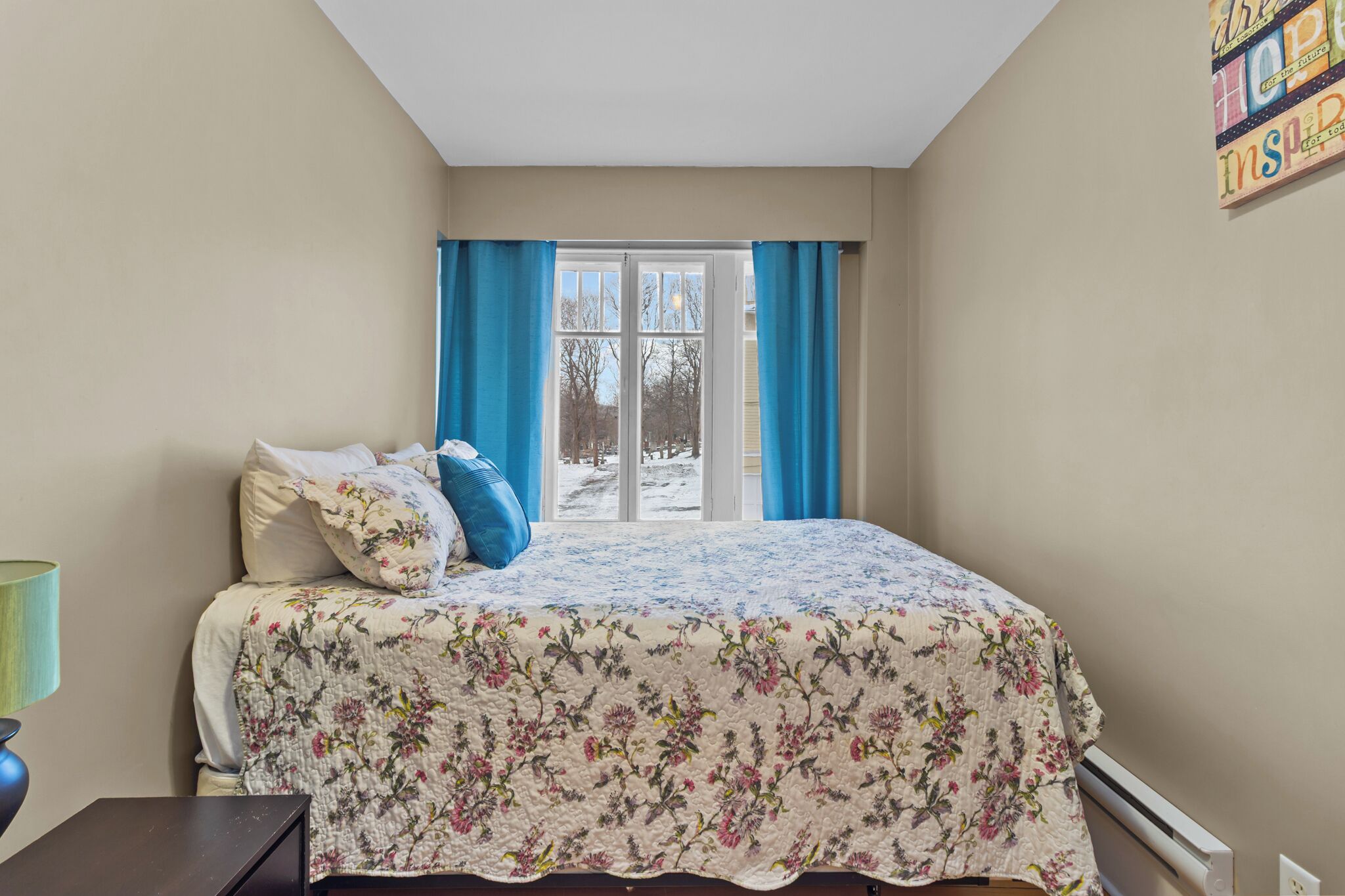 A bedroom with a bed, a window with curtains, and a wall with a motivational poster.