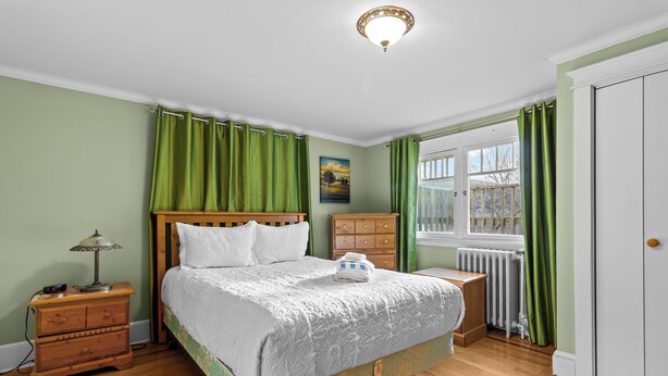 A bedroom with a wooden bed, green curtains, a nightstand, a lamp, and a window with a view. - Midstream Manor 1BR Parking WiFi (St. John's)