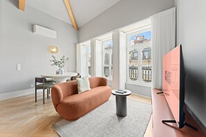 Una sala moderna con un sofá, una mesita, un televisor y una ventana con vistas a edificios.