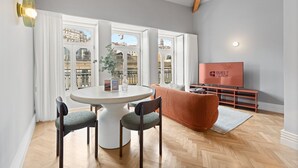 A modern living room with a round dining table, two chairs, a sofa, a TV, and a view of a cityscape through large windows.
