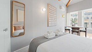 A hotel room with a large bed, a mirror, a wall-mounted artwork, a table with chairs, and a window with a view of buildings.