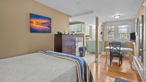 A bedroom with a bed, a painting, a dresser, and a view to a kitchen and dining area. - Chic Midstream Studio WiFi Parking (St. John's)