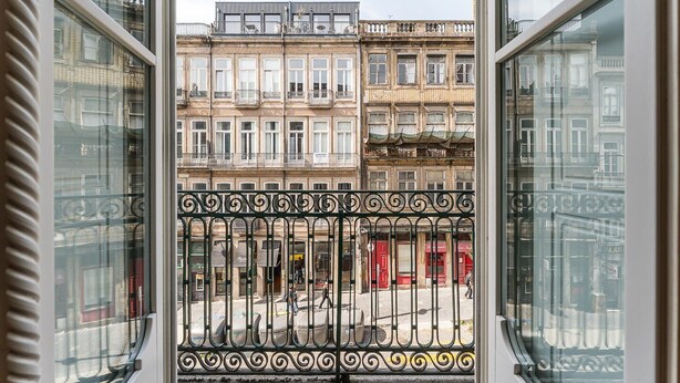 Blick aus einem Fenster mit aufwendigen schwarzen Metallgeländern auf eine Straße mit mehrstöckigen Gebäuden und einem roten Schaufenster.