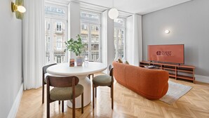 Ein modernes Wohnzimmer mit einem runden Esstisch, zwei Stühlen, einer Couch, einem Fernseher und Blick auf Gebäude durch große Fenster.