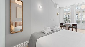 Ein modernes Hotelzimmer mit einem großen Bett, einem Spiegel, einem Essbereich mit Stühlen und Blick auf die Stadt durch große Fenster.