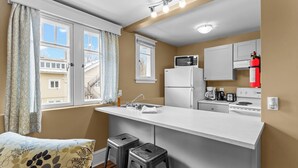 Une cuisine compacte avec un plan de travail blanc, un four à micro-ondes, un réfrigérateur et un évier. On y trouve deux tabourets et une fenêtre avec des rideaux.