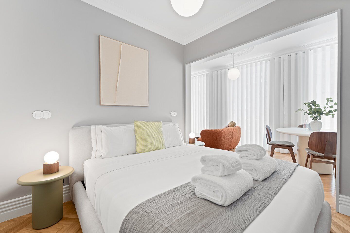 A modern hotel room with a large bed, a bedside table with a lamp, a chair, a small table with a plant, and a window with sheer curtains.