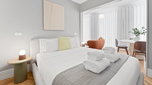 Ein modernes Hotelzimmer mit einem großen Bett, einem Nachttisch mit Lampe, einem Sessel, einem kleinen Tisch mit Pflanze und einem Fenster mit durchsichtigen Vorhängen.