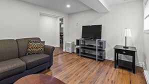 A modern living room with a grey sofa, a flat-screen TV on a black stand, a wooden coffee table, and a small black side table with a lamp. - Stylish 2BR WiFi Parking MUN Walk (St. John's)