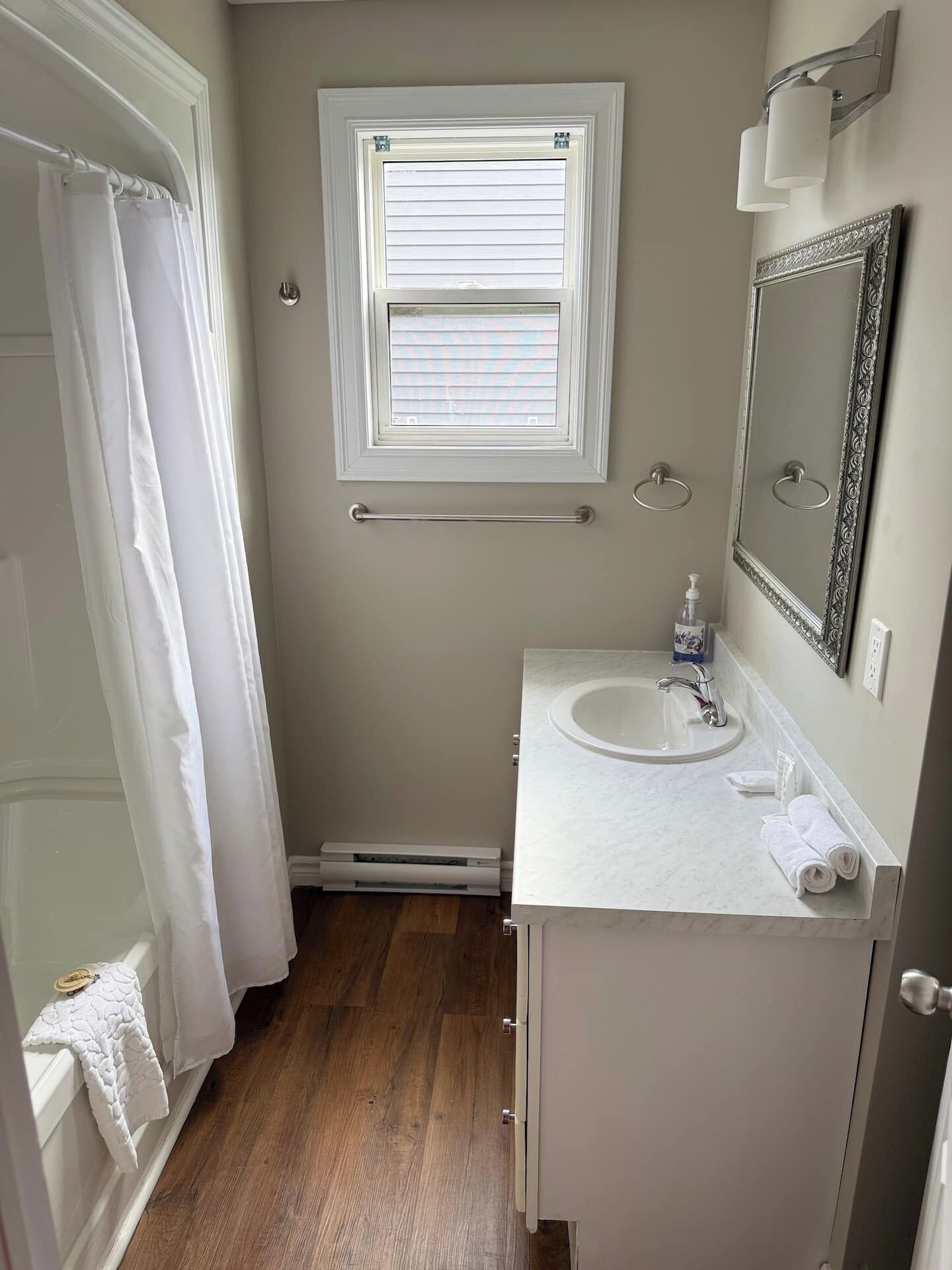 Un baño con un lavamanos blanco, un espejo, una ventana con persianas, un perchero para toallas y una cortina de ducha.