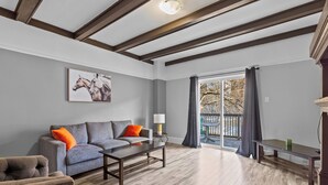 Un salon spacieux avec un canapé gris, une table basse et un plancher en bois. Une grande fenêtre offre une vue sur les arbres, et il y a un balcon.