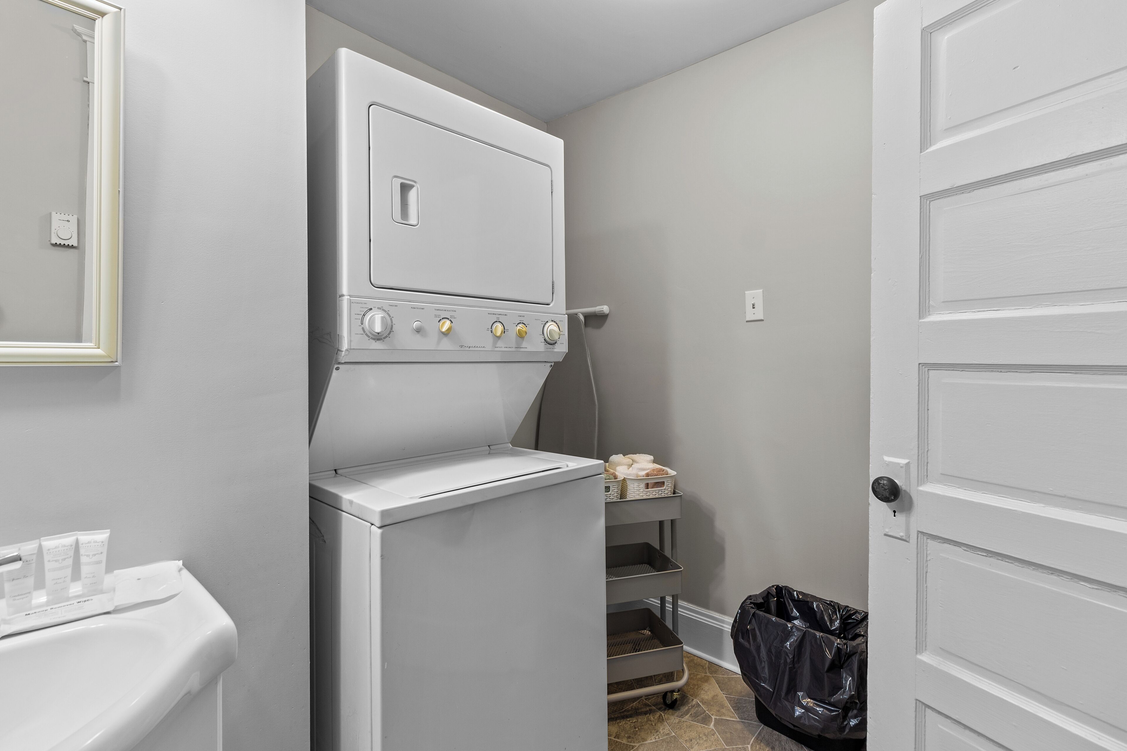 A compact laundry room with a washer and dryer, a small shelf with baskets, and a trash bin.